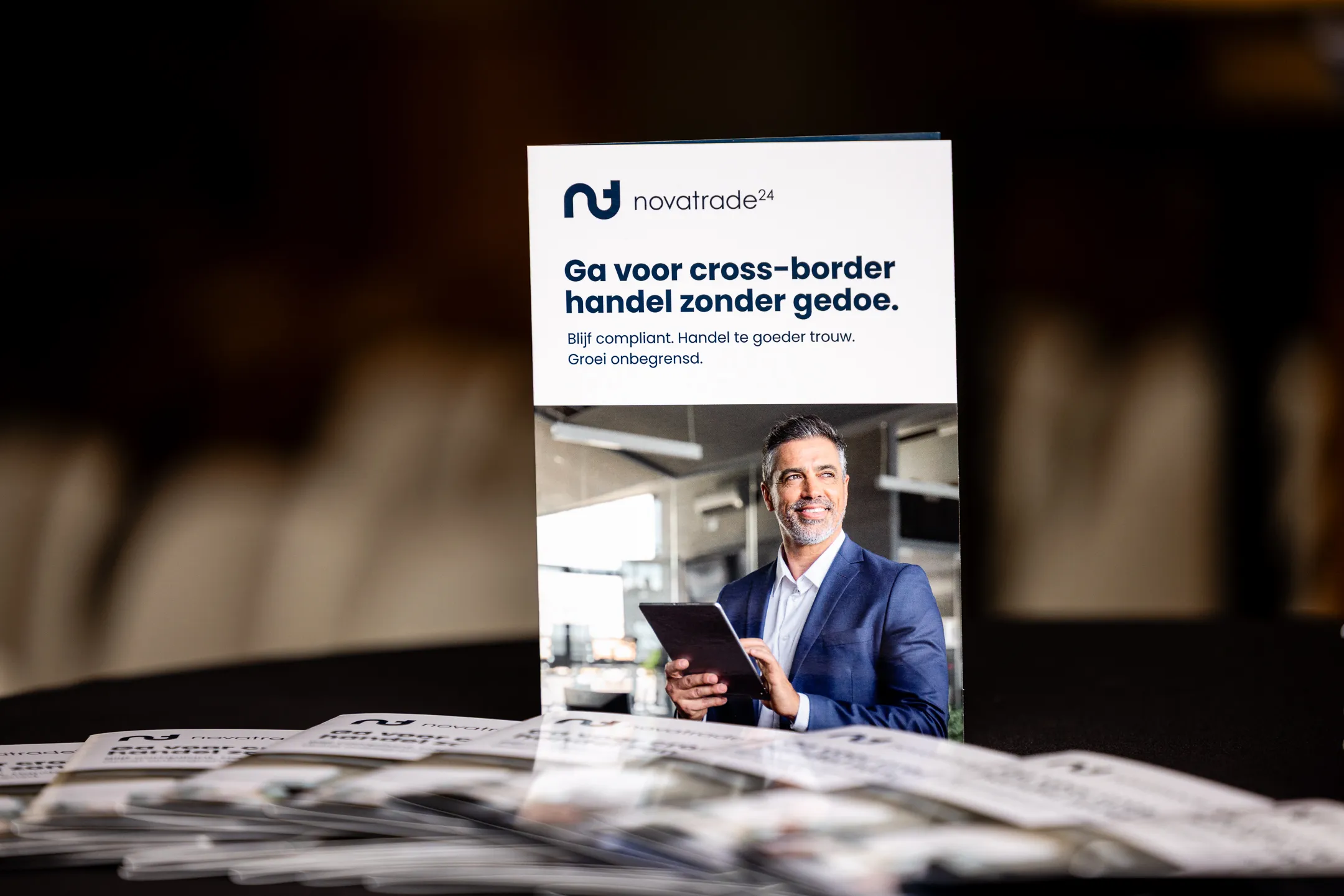 A Novatrade24 brochure standing on a table, with more brochures spread out in front of it