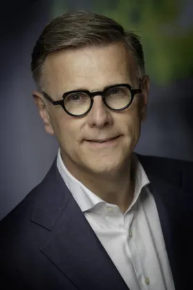 Portrait of Michel van Roon with round dark glasses, in a navy jacket and open-collar white shirt