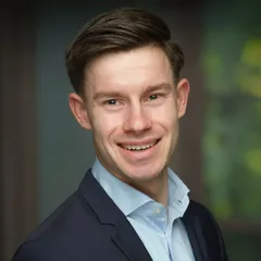 Portret van Tom Hessing, glimlachend, in een donker colbert en lichtblauw overhemd